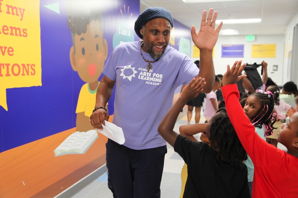 A teacher, smiling as he walks down the hallway, holds his hand up for high fives. A line of students are walking on the opposite side with hands up and ready.
