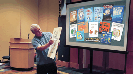 The artist, Tim Young, holds a dry-erase board while he sketches. He is standing in front of a large projection displaying his illustrated book covers.