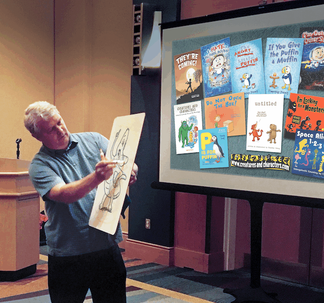 The artist, Tim Young, holds a dry-erase board while he sketches. He is standing in front of a large projection displaying his illustrated book covers.