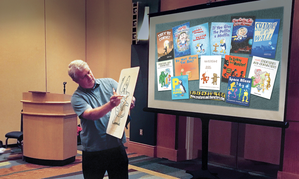 The artist, Tim Young, holds a dry-erase board while he sketches. He is standing in front of a large projection displaying his illustrated book covers.