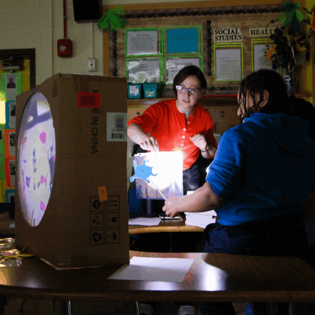 A teaching artist and students behind a shadow theatre constructed out of a cardboard box with a round screen. A bright light shines from behind.