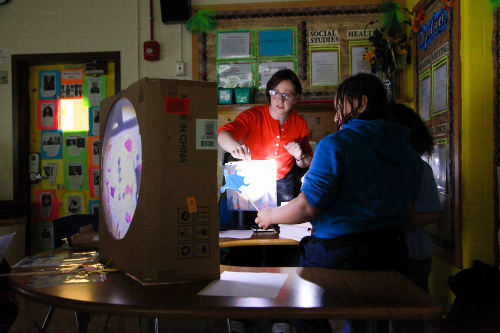 A teaching artist and students behind a shadow theatre constructed out of a cardboard box with a round screen. A bright light shines from behind.