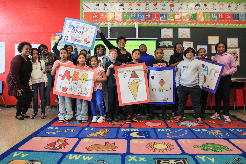 Students pose with their teacher and teaching artist Maria Weaver, holding the posters they created in small groups.