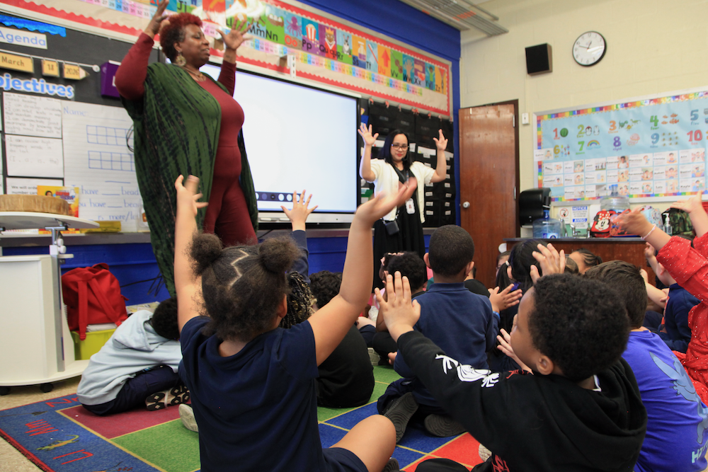 A teacher and teaching artist lead the seated Kindergarteners in movement and rhythm.