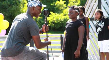 Femi the Drifish adjusts a microphone for young performers on an outdoor stage.