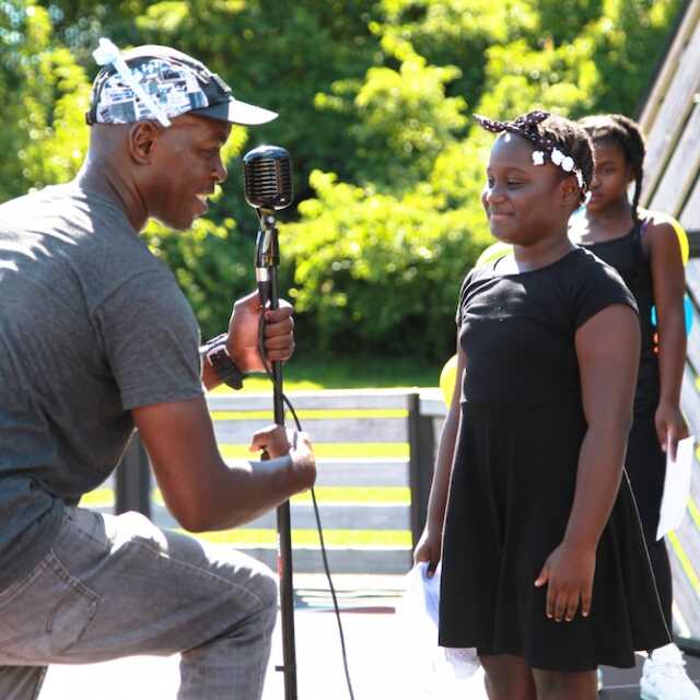 Femi the Drifish adjusts a microphone for young performers on an outdoor stage.