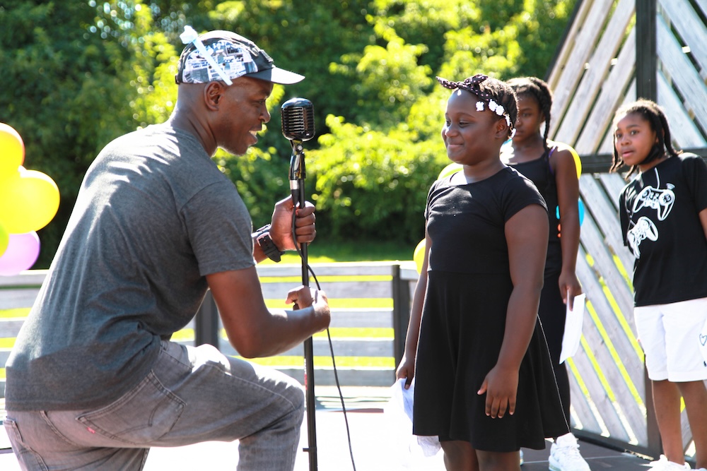 Femi the Drifish adjusts a microphone for young performers on an outdoor stage.