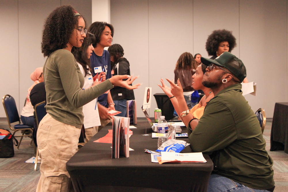 High school students standing to talk with representatives from arts and cultural organizations who are seated at a long table.