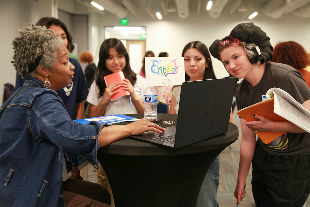 Students surround a representative from Empact Art and look curiously at the presenter's laptop screen.