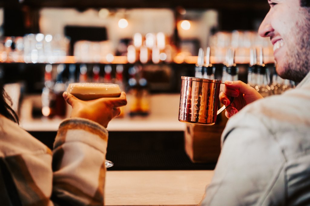 Two people enjoying a cozy moment at a bar, each holding a warm beverage in a copper mug and a glass, with a blurred background of bottles and glasses in Williamson County, Tennessee.