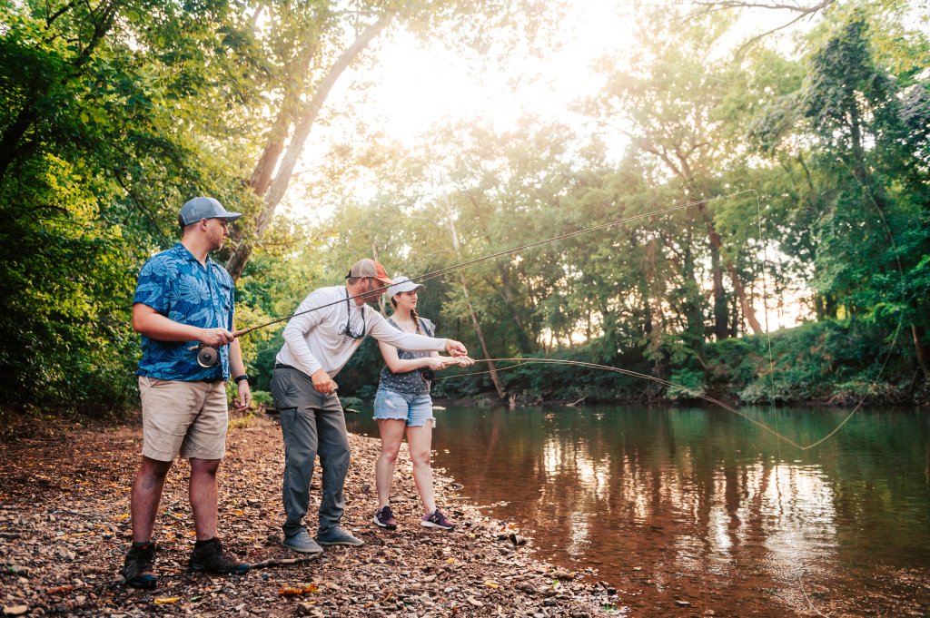 Three people are fishing on a serene riverbank, surrounded by lush greenery and dappled sunlight in Williamson County, Tennessee.