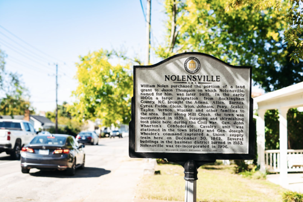 A historical marker sign for Nolensville, Tennessee, stands on a grassy area beside a road with cars passing by in Williamson County, Tennessee.