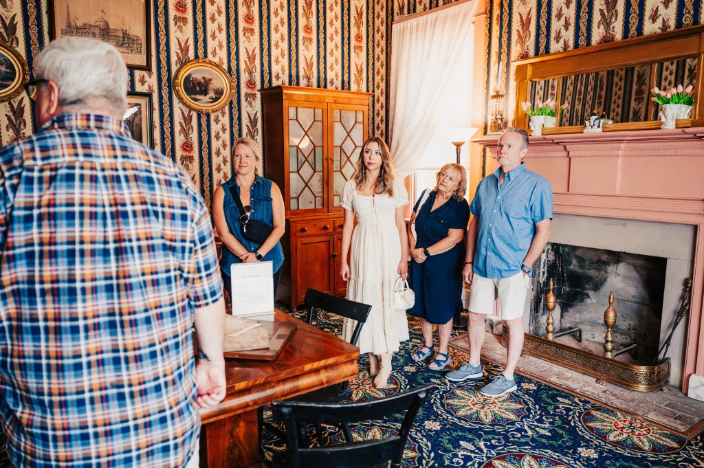 A group of people, including an elderly man in a plaid shirt, are gathered in a room with ornate wallpaper and a fireplace, listening to a tour guide in Williamson County, Tennessee.