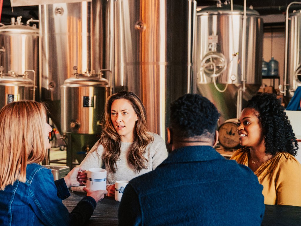 Four people are gathered around a table in a brewery, holding coffee mugs and engaged in conversation, with large brewing tanks in the background in Williamson County, Tennessee.
