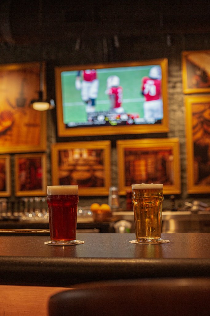 Two glasses of beer, one dark and one light, sit on a bar counter with a football game playing on a large TV in the background in Williamson County, Tennessee.