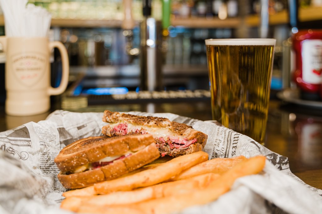 A mouthwatering Reuben sandwich, cut in half and served on toasted rye bread with melted cheese and corned beef, accompanied by a tall glass of golden beer and a side of crispy french fries in Williamson County, Tennessee.