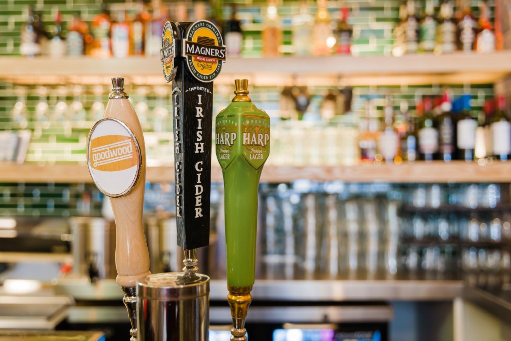 Three beer taps with labels for Goodwood, Magners Irish Cider, and Harp Lager, set against a backdrop of a well-stocked bar with green-tiled shelves in Williamson County, Tennessee.