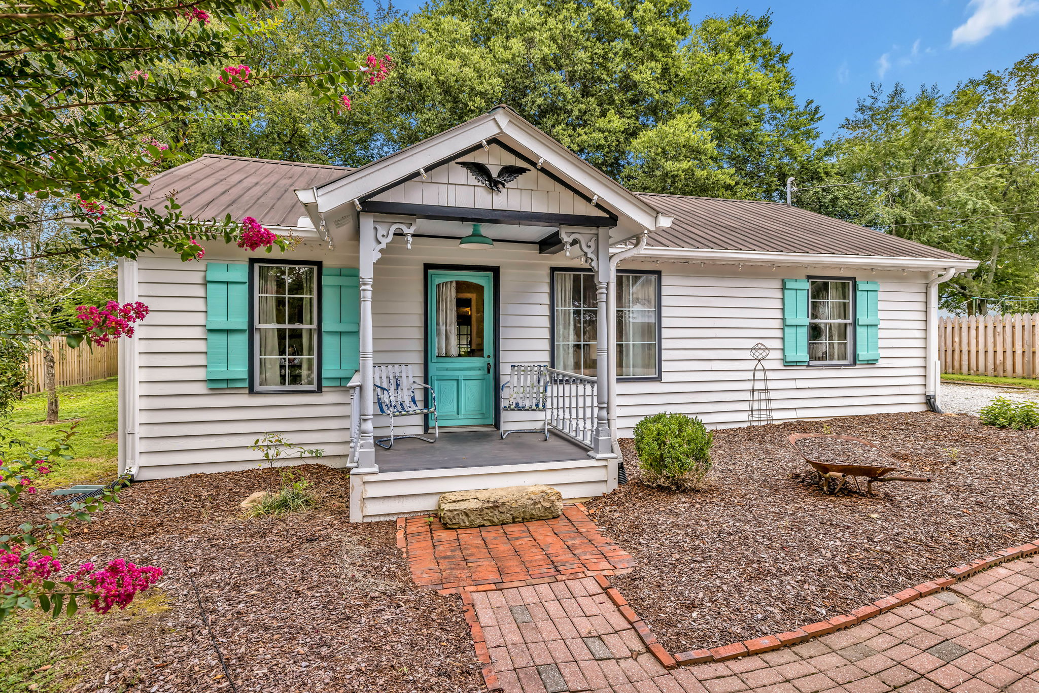 A charming white cottage with turquoise shutters and a welcoming front porch, nestled among lush greenery and colorful flowers in Williamson County, Tennessee.