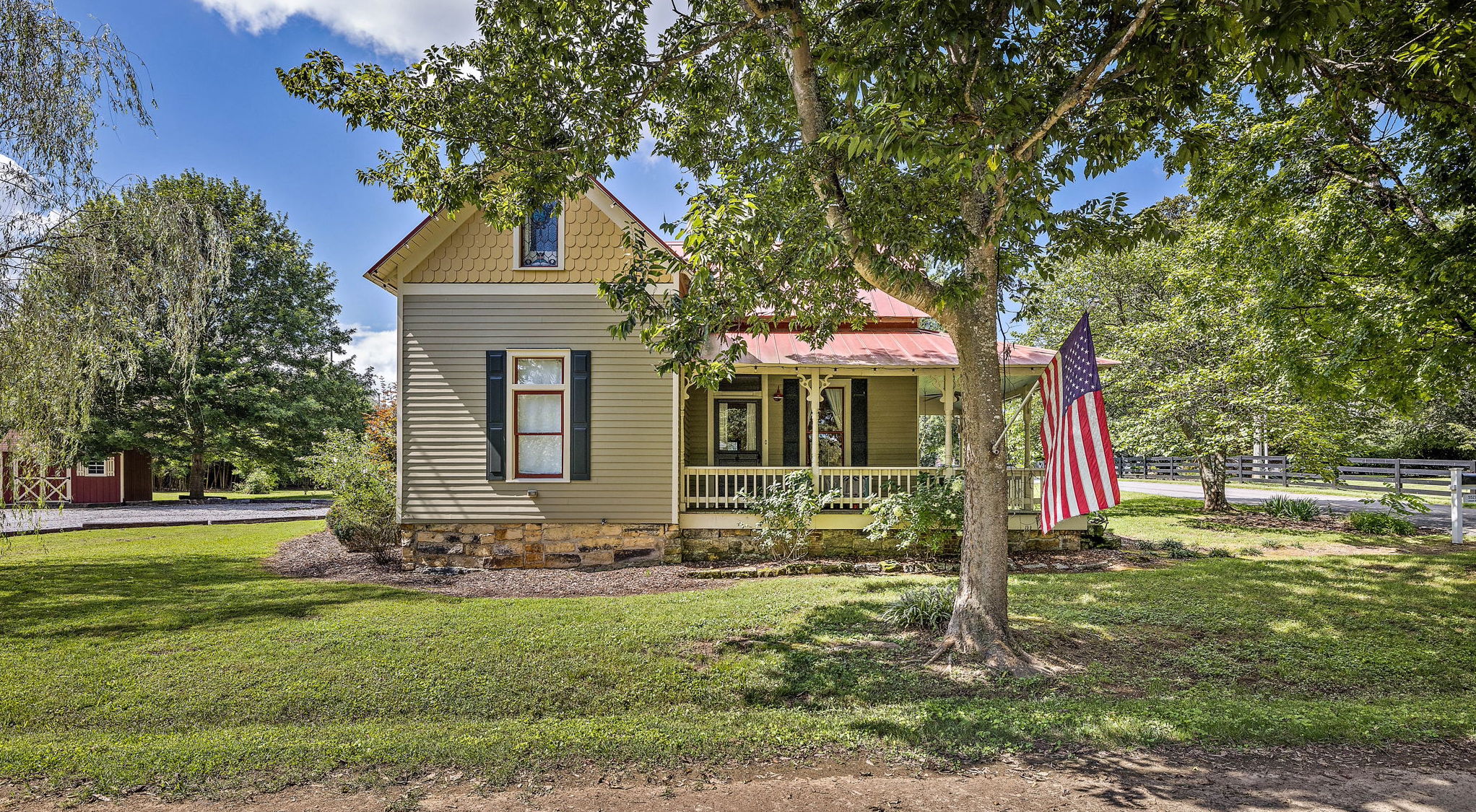 A charming two-story farmhouse with a wrap-around porch and an American flag stands amidst lush greenery and tall trees in Williamson County, Tennessee.