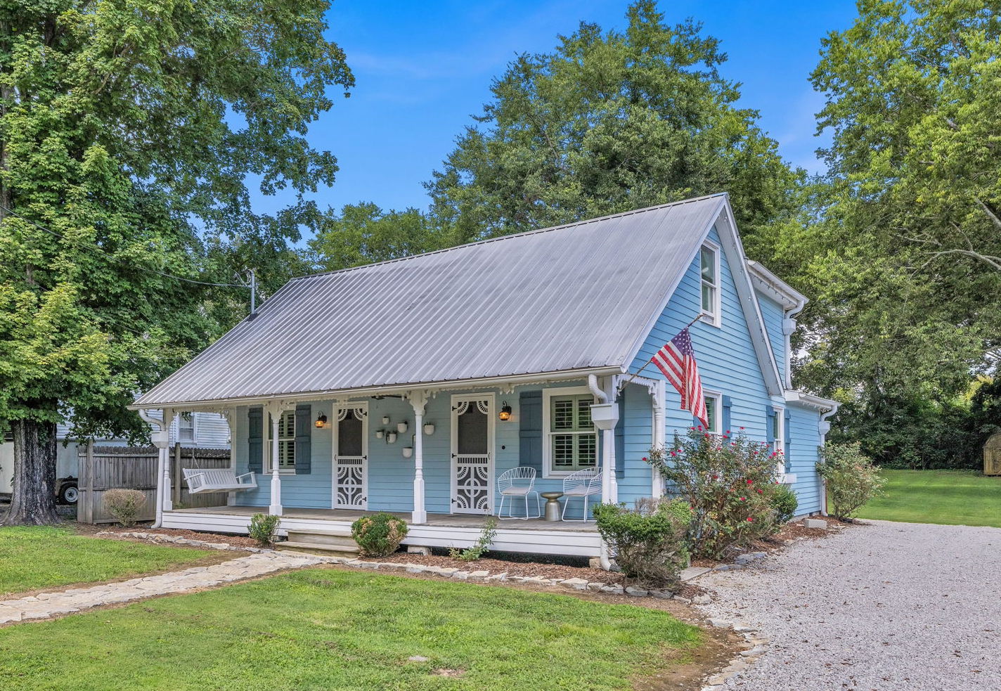 A charming blue cottage with a metal roof, white trim, and a welcoming front porch adorned with American flags and rocking chairs, set against a backdrop of lush green trees and a clear blue sky in Williamson County, Tennessee.