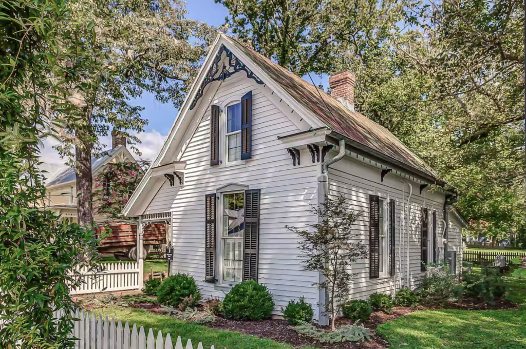 A charming white Victorian cottage with black shutters and a wraparound porch, nestled among lush greenery and a white picket fence in Williamson County, Tennessee.