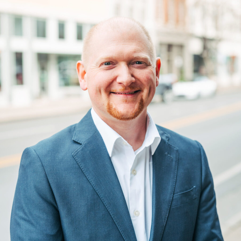 A smiling man with a beard wearing a blue suit stands on a city street in Williamson County, Tennessee.
