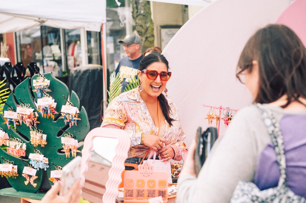 A woman with sunglasses and a floral shirt smiles broadly at a market stall displaying colorful jewelry and accessories in Williamson County, Tennessee.