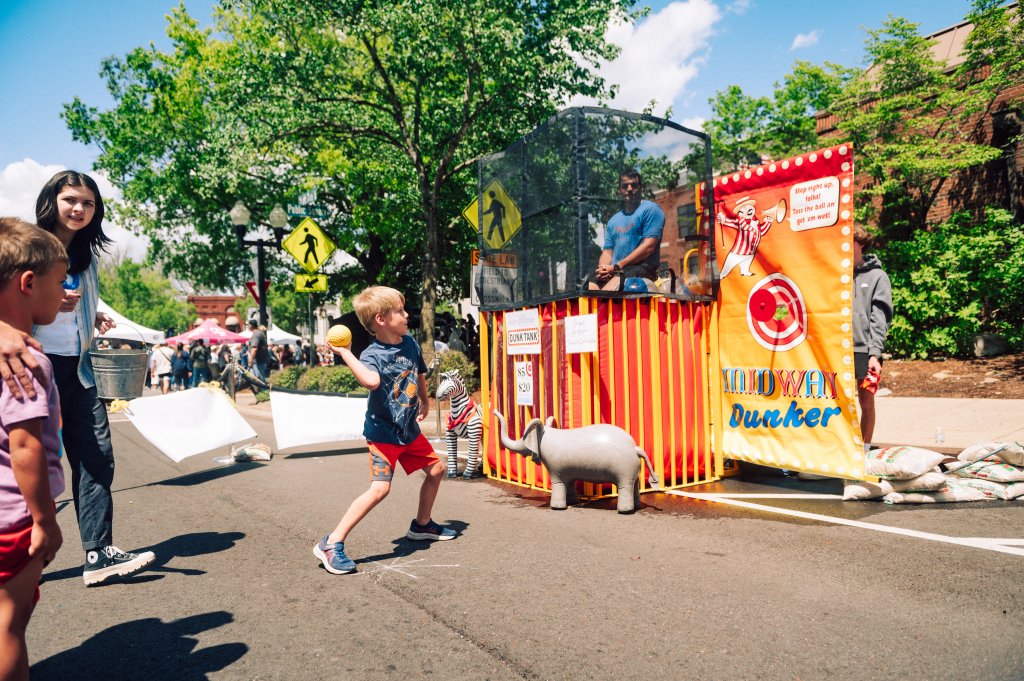 A young boy joyfully throws a ball at a colorful dunk tank during a lively outdoor festival, with a crowd of onlookers cheering him on in Williamson County, Tennessee.