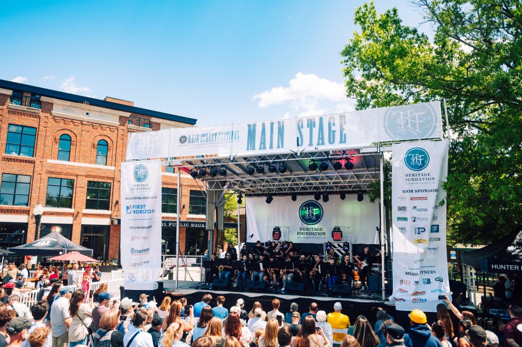 A lively outdoor music festival with a large crowd gathered in front of a stage adorned with banners and logos, set against a backdrop of a historic brick building and lush green trees in Williamson County, Tennessee.