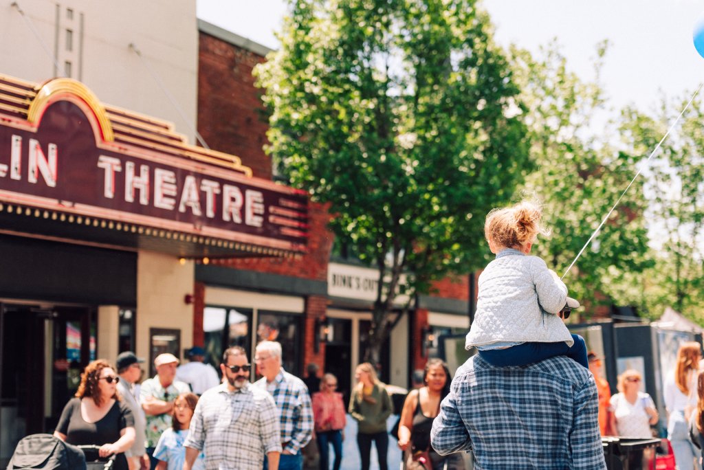 A lively street scene in front of the historic Elgin Theatre, where a child rides on a man's shoulders, holding a blue balloon string in Williamson County, Tennessee.
