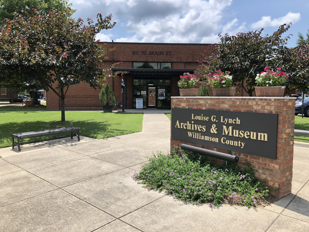 A quaint brick building with a sign reading 'Louise G. Lynch Archives & Museum, Williamson County' surrounded by lush greenery and colorful flowers in Williamson County, Tennessee.