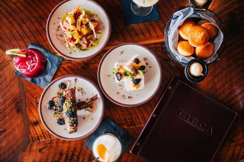 A variety of dishes including a colorful salad, grilled lamb chops with blackberries, a dessert with berries and nuts, and a basket of biscuits, accompanied by cocktails and a menu book in Williamson County, Tennessee.