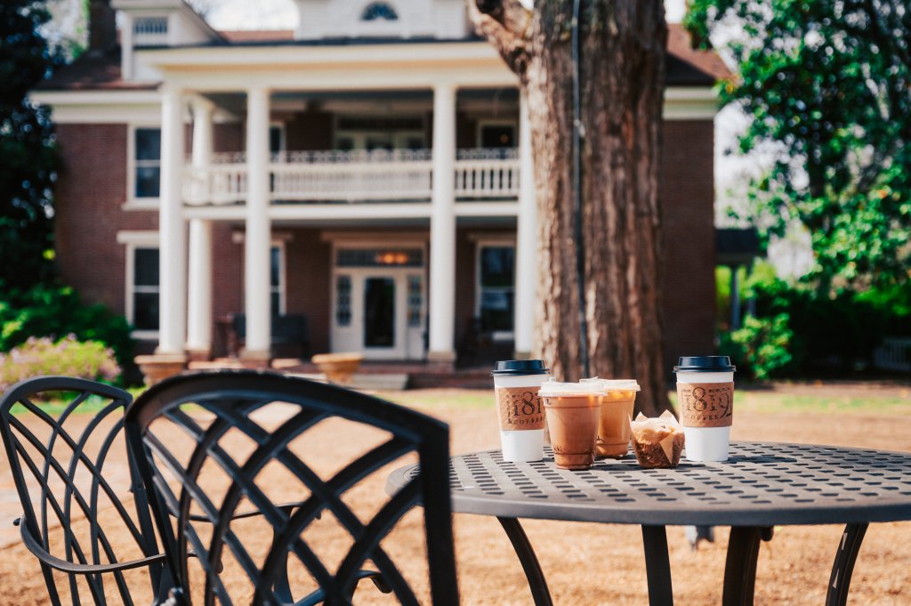 Three coffee cups and a muffin sit on a metal table in a garden, with a historic mansion and a large tree in the background in Williamson County, Tennessee.