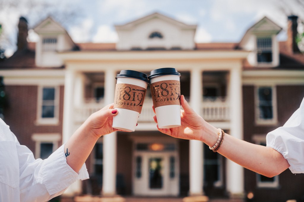 Two people cheerfully clink disposable coffee cups in front of a grand, historic mansion with a wide porch and columns in Williamson County, Tennessee.