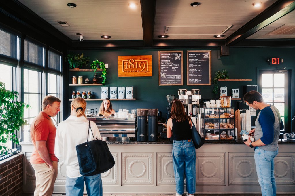 Patrons gather at the counter of a cozy coffee shop with dark walls and large windows, ordering drinks and pastries from the baristas in Williamson County, Tennessee.