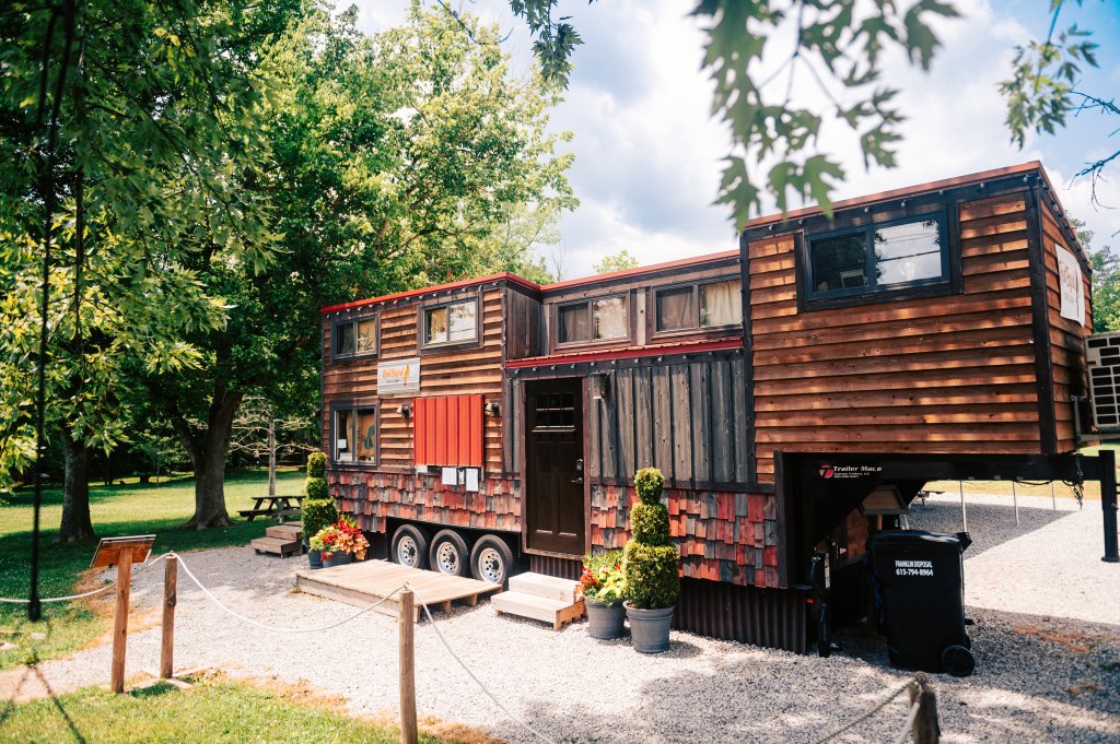 A rustic tiny house on wheels with a red roof is parked in a wooded area in Williamson County, Tennessee.