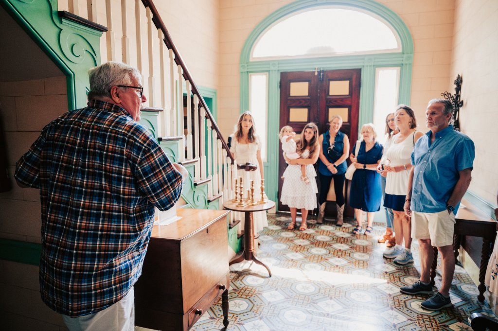 A group of people, including an elderly man in a plaid shirt, gather in a beautifully decorated hallway with green staircases and a tiled floor, listening attentively to a tour guide in Williamson County, Tennessee.