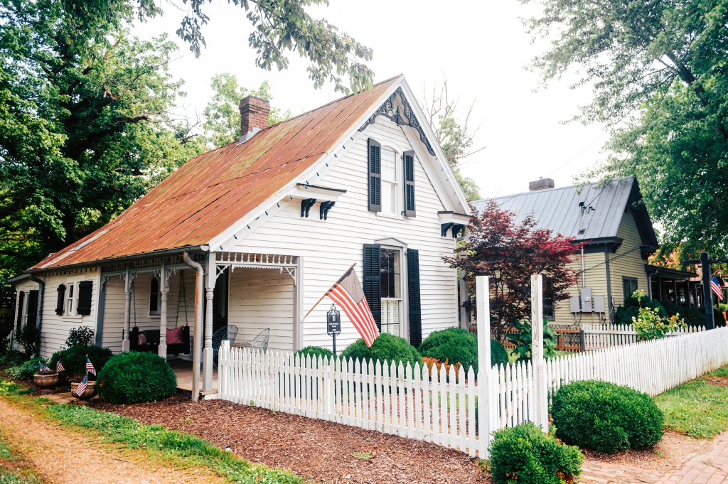 A charming white house with a red roof and black shutters is nestled among lush greenery, featuring a welcoming porch with a swing and an American flag flying proudly in Williamson County, Tennessee.