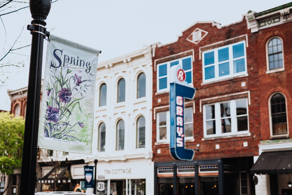 A charming downtown street scene with a 'Spring' banner featuring purple flowers hanging from a lamppost, set against a backdrop of historic brick and white buildings with large windows and vintage signage in Williamson County, Tennessee.
