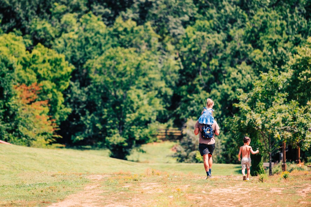 A man carries a child on his shoulders while walking with another child in a lush, green forest setting in Williamson County, Tennessee.
