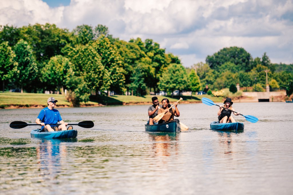 Three people are kayaking on a serene lake surrounded by lush green trees and a clear blue sky in Williamson County, Tennessee.