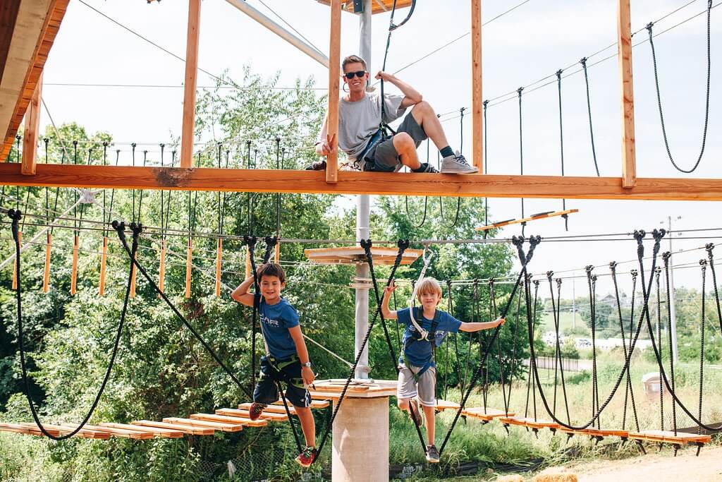 Three children and an adult are enjoying an aerial ropes course, with the children suspended on ropes and the adult sitting on a wooden platform above in Williamson County, Tennessee.