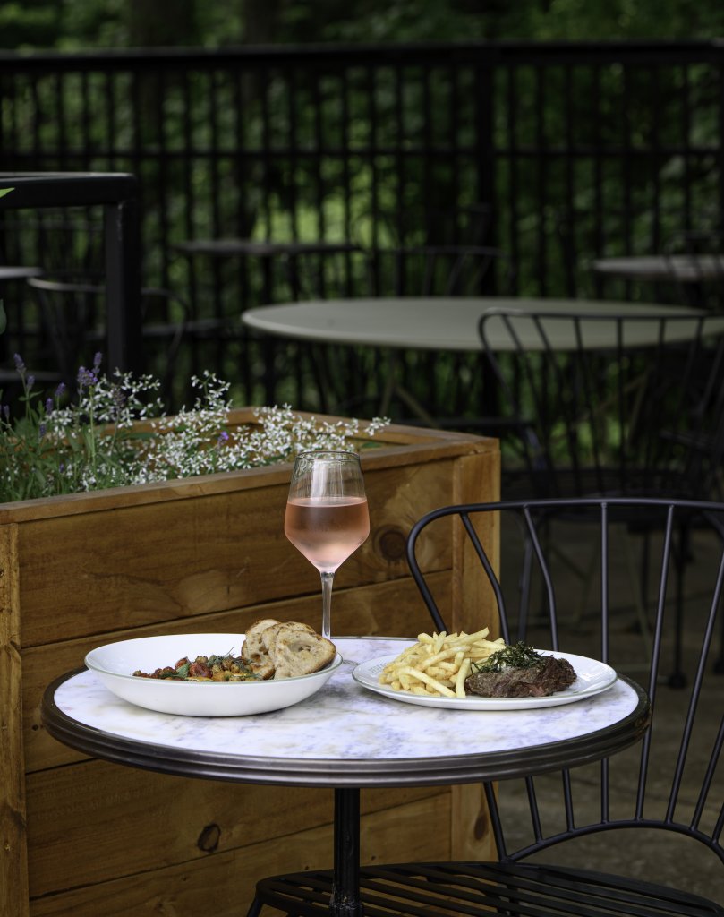 A romantic outdoor dining setup with a glass of rosé wine, a plate of steak and fries, and a bowl of salad on a small round table, surrounded by lush greenery and blooming flowers in Williamson County, Tennessee.