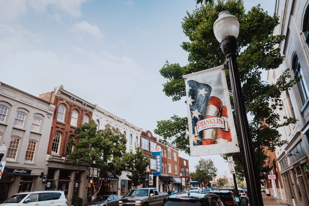 A bustling downtown street with colorful buildings, parked cars, and an American flag banner in Williamson County, Tennessee.