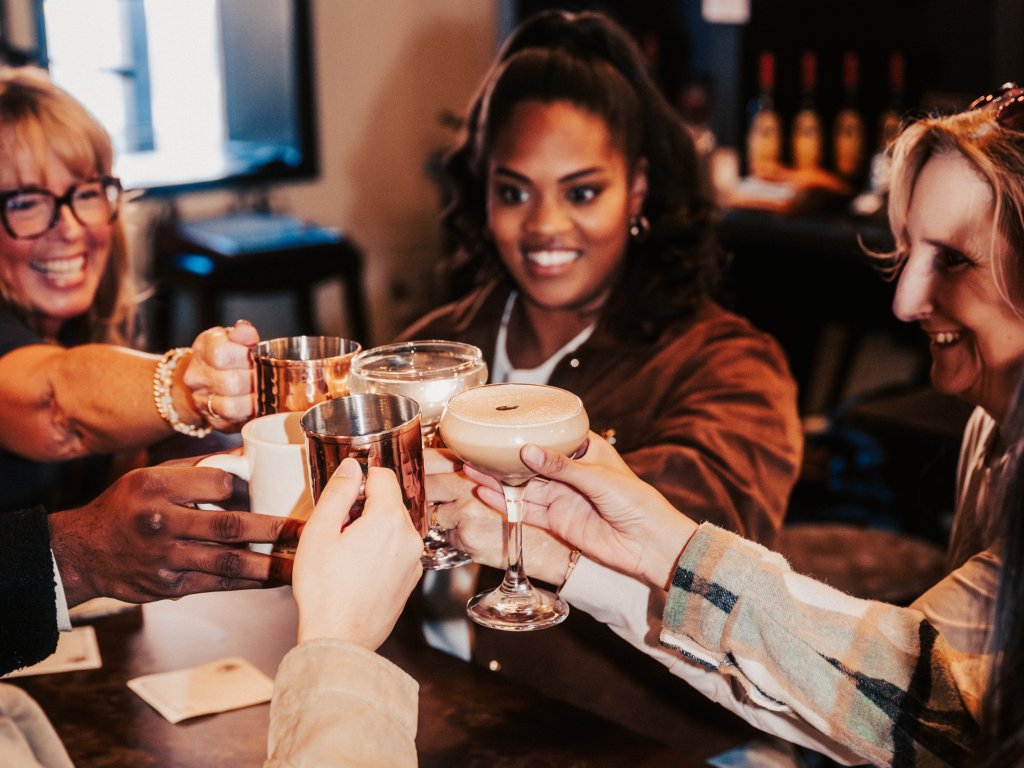 6 of Franklin's Best Coffees & Cocktails to Drink in the Fall Season A diverse group of friends cheers with cocktails and mugs in a dimly lit bar in Williamson County, Tennessee.