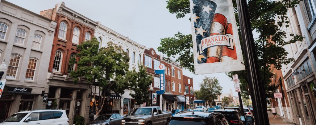 A bustling downtown street with historic buildings, parked cars, and a patriotic banner in Williamson County, Tennessee.