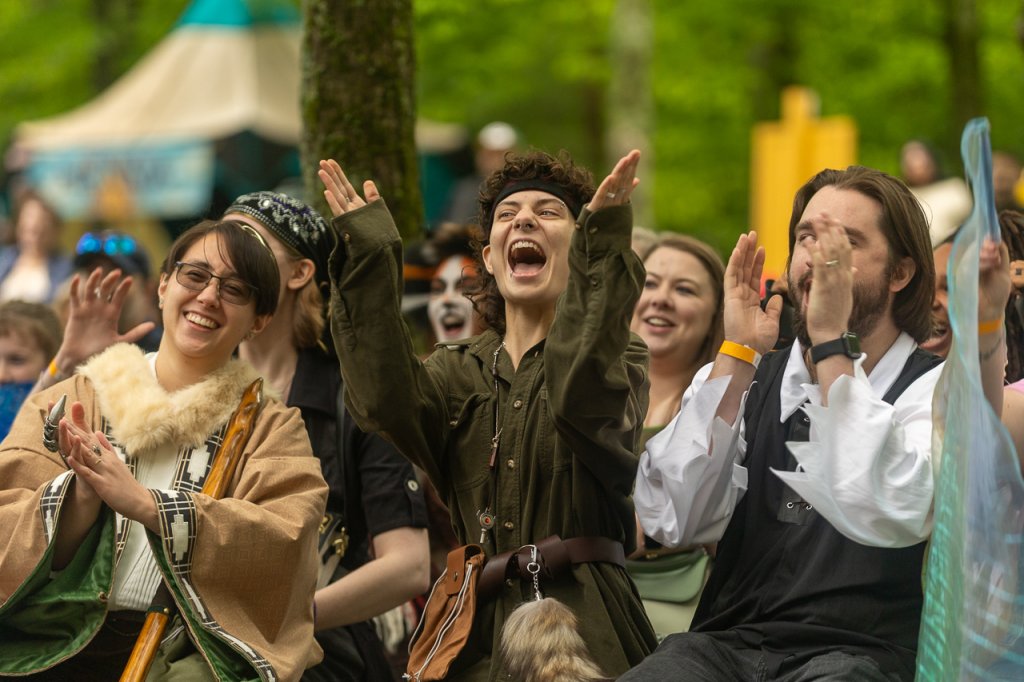 A diverse group of people dressed in various costumes, including a woman in a fur-trimmed cloak and a man in a green military-style jacket, are cheering and clapping enthusiastically in a forest setting in Williamson County, Tennessee.