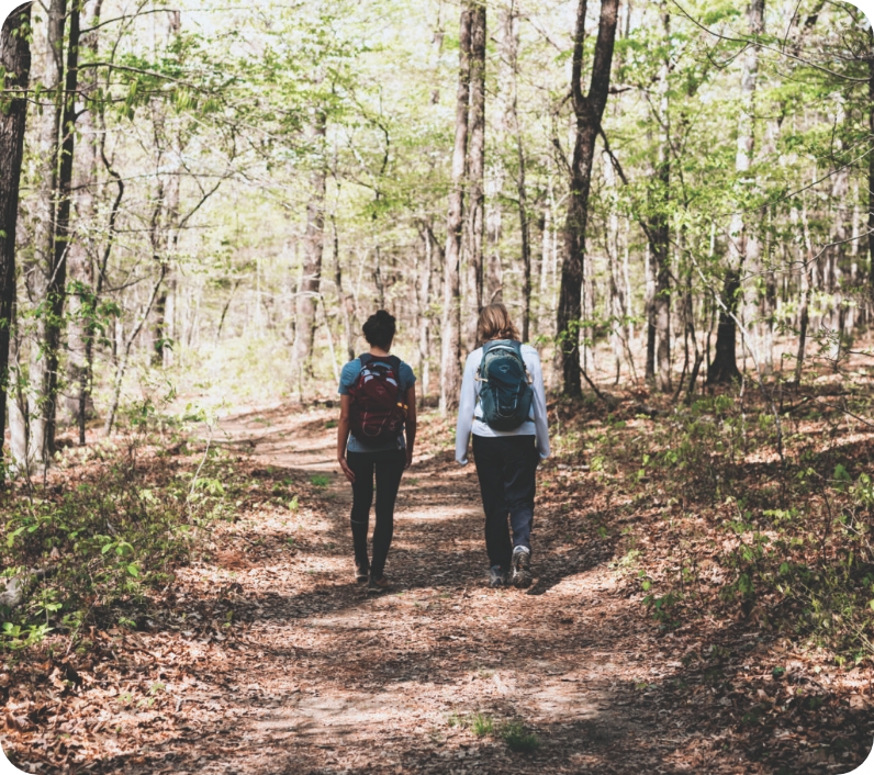 Two hikers with backpacks walk along a forest trail lined with trees and fallen leaves in Williamson County, Tennessee.