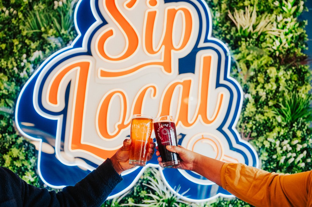 Two people clinking glasses of beer in front of a 'Sip Local' neon sign in Williamson County, Tennessee.