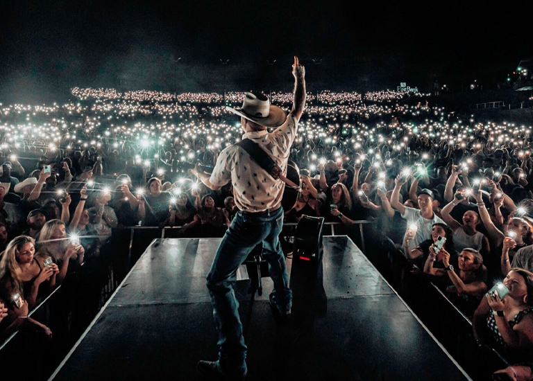 A country music performer in a cowboy hat stands on stage, arm raised, facing a sea of fans with lit phones in Williamson County, Tennessee.